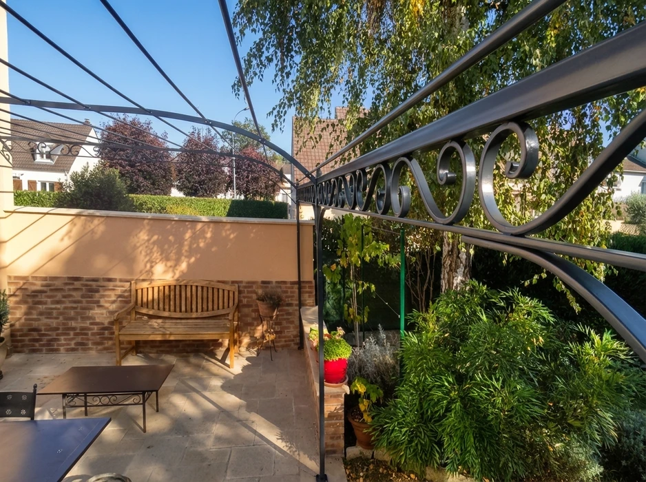 Vue d'une marquise en fer forgé installée au-dessus d'une entrée résidentielle ou d'une terrasse, présentant une structure métallique élaborée avec volutes décoratives et motifs floraux ou géométriques typiques de la ferronnerie d'art, en noir ou gris, créant un élégant auvent ajouré sur un bâtiment de prestige ou une maison ancienne, avec un contraste clair entre la structure forgée et le mur ou la fenêtre en arrière-plan, photographiée de jour sous un bon éclairage.